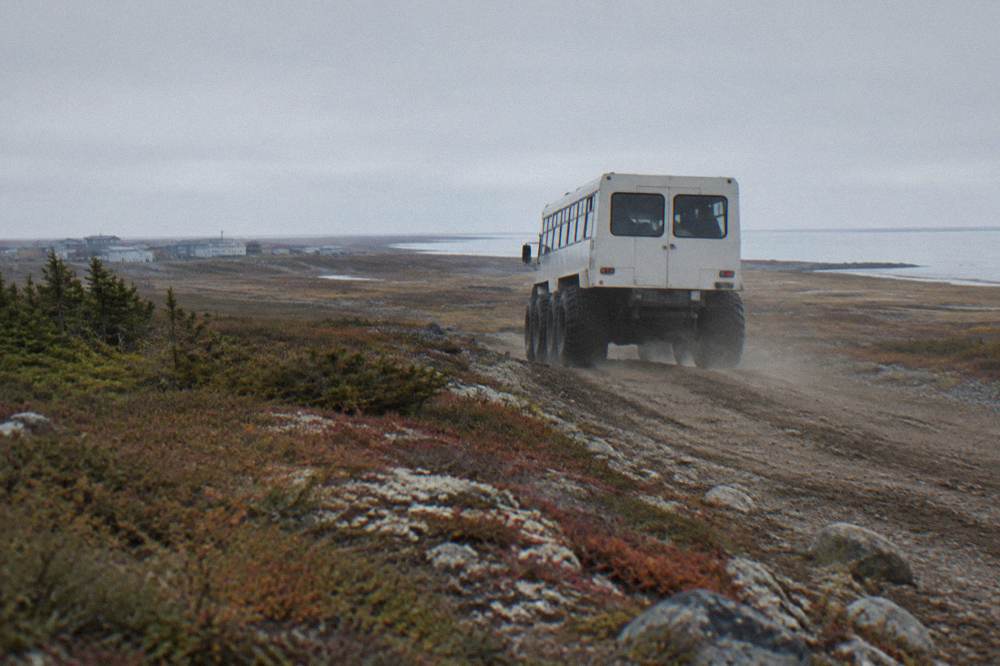 A tall tundra vehicle travelling on a rough track across wide open tundra toward a distant shoreline