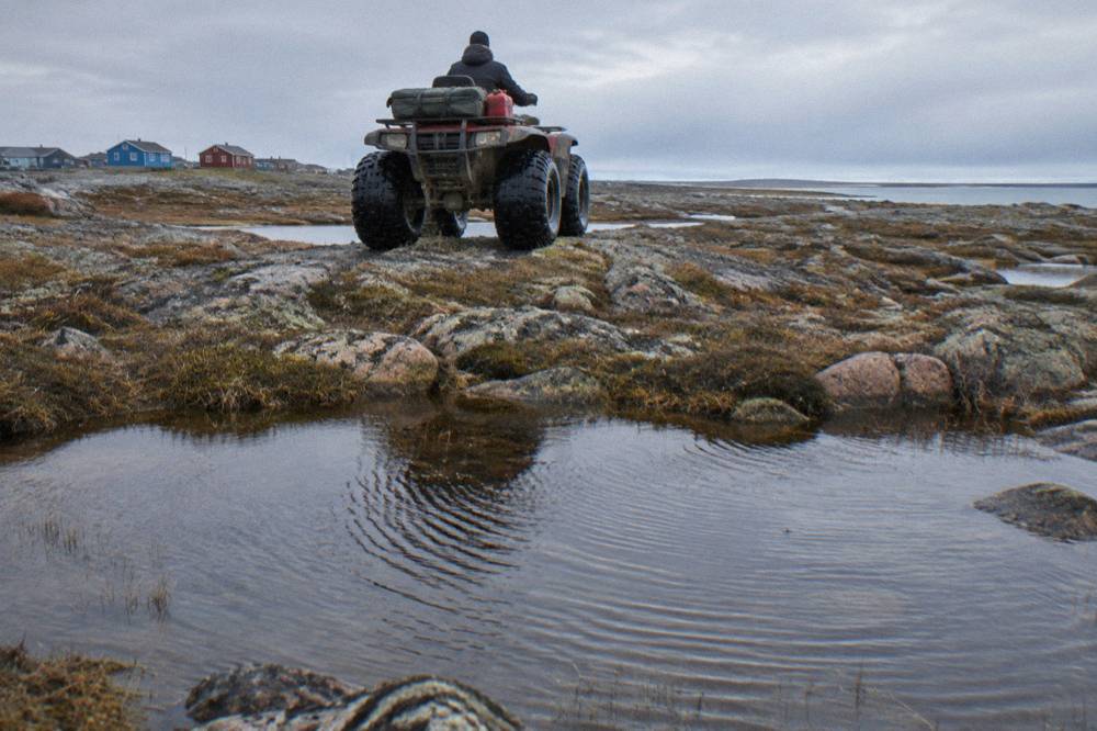 A raised tundra vehicle with large tires crossing open subarctic terrain dotted with ponds
