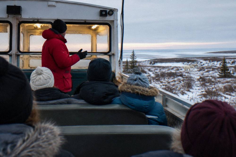 A guide in a red parka speaking to travelers aboard a raised tundra vehicle deck