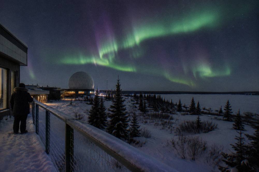 A green and purple aurora arc stretching across a dark night sky above snowy tundra