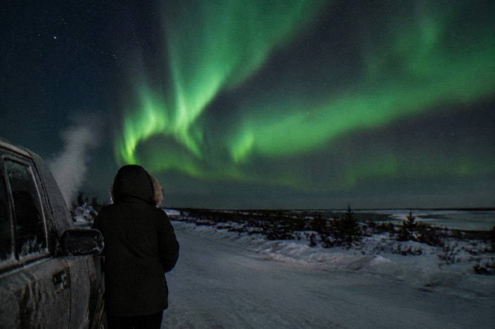A person in a heavy winter coat watching a bright aurora display from beside a snow-covered vehicle
