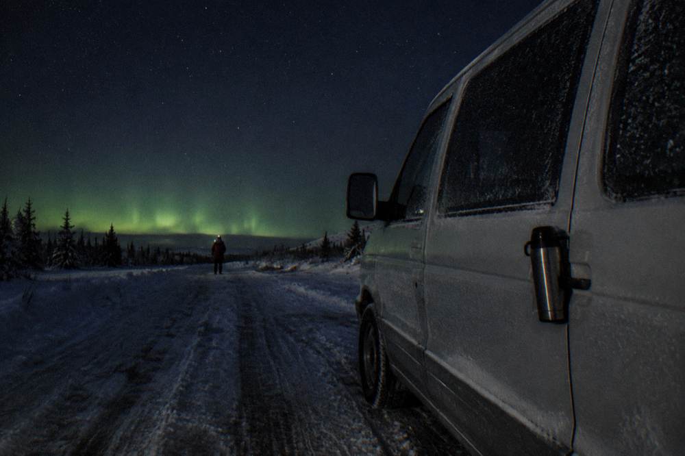 A heated tour vehicle parked on a dark snowy road under a starry sky with faint green aurora on the horizon