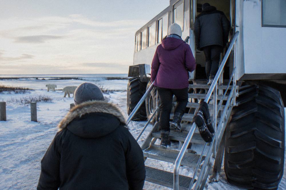 A large wheeled tundra vehicle at a gravel launch site with guests boarding via a raised staircase