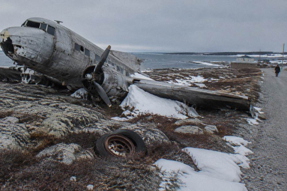 A weathered propeller aircraft wreck resting on rocks beside a coastal gravel road