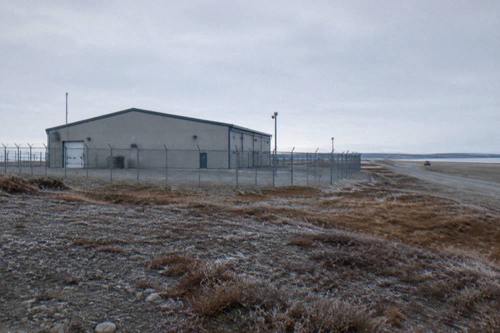 A large industrial building with a tall fence near an airport runway in a flat northern landscape