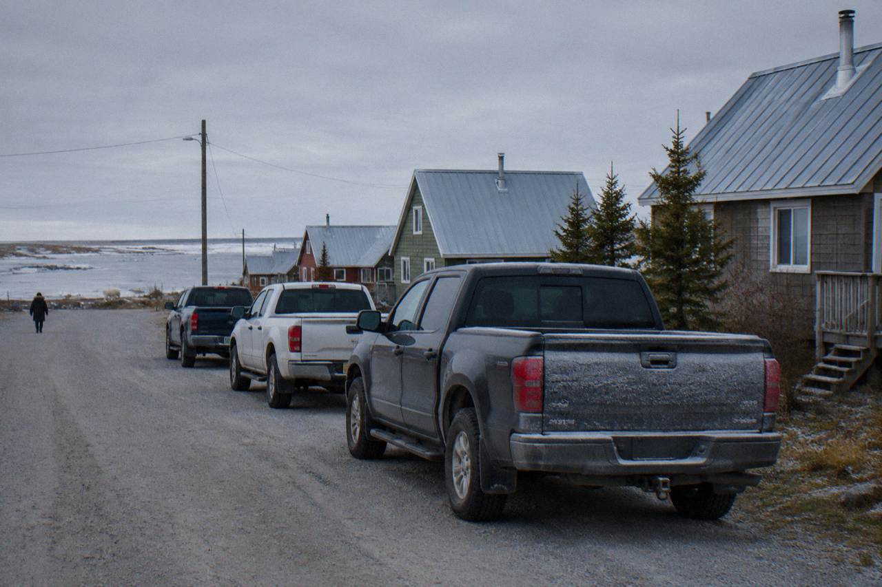 A row of pickup trucks parked on a quiet gravel street in a small northern town under a wide grey sky