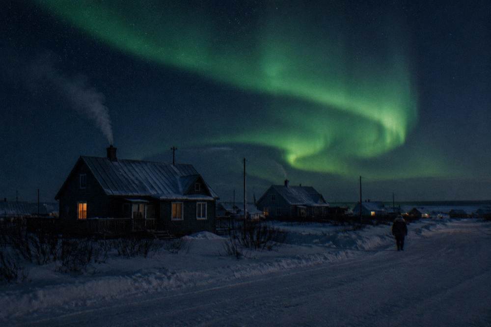 Green aurora light curving above a dark snowy landscape with low buildings silhouetted