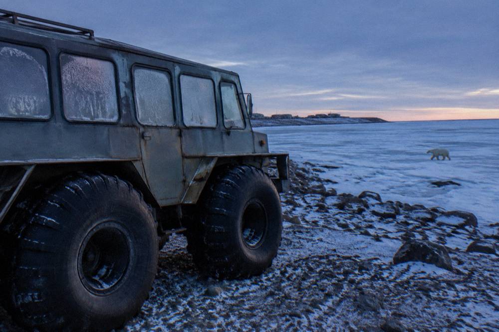 A purpose-built raised tundra vehicle with huge tires parked on open rocky ground near a frozen bay