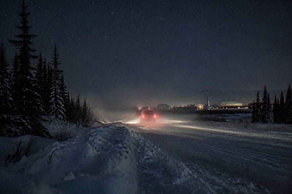 A headlights-on vehicle traveling a snow-packed road at night under a starry sky