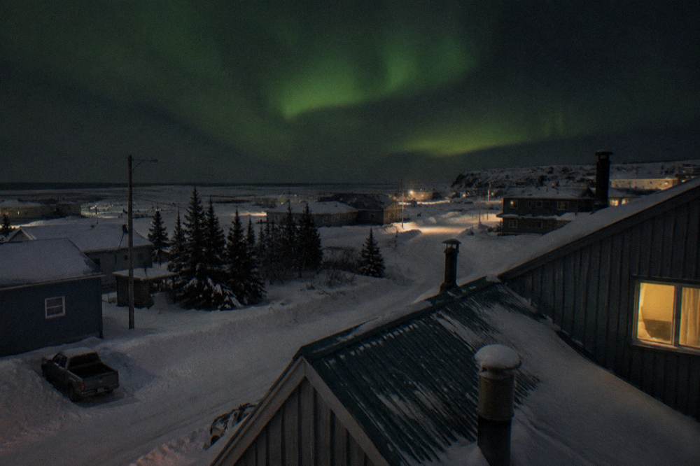 A small northern town at night with snow-covered buildings and a green glow in the sky above