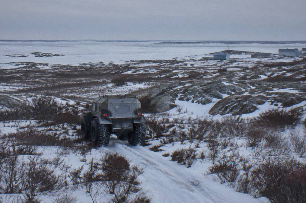 A raised tundra vehicle with large tires crossing flat open tundra dotted with patches of snow