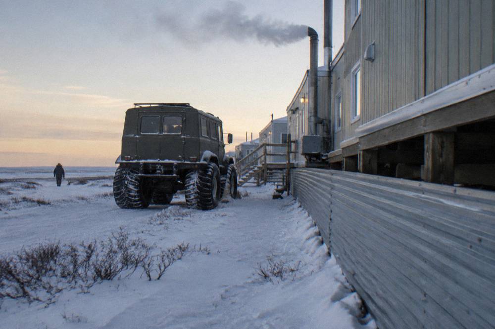 An elevated tundra vehicle with large tires parked beside a mobile lodge complex on flat snow-covered tundra