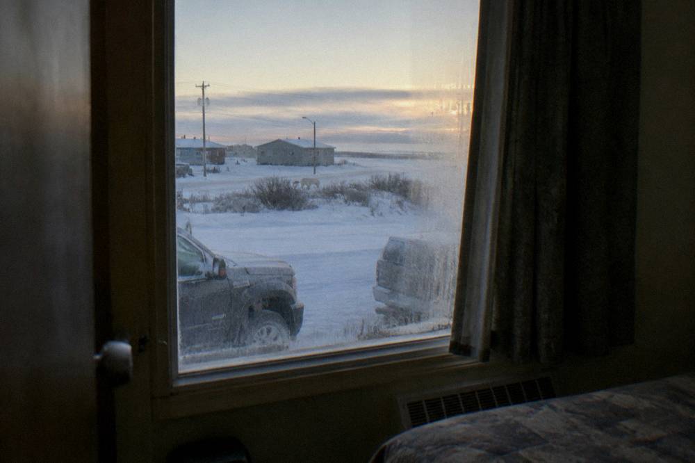 A hotel room window in Churchill overlooking a snow-covered street with parked vehicles under early morning light