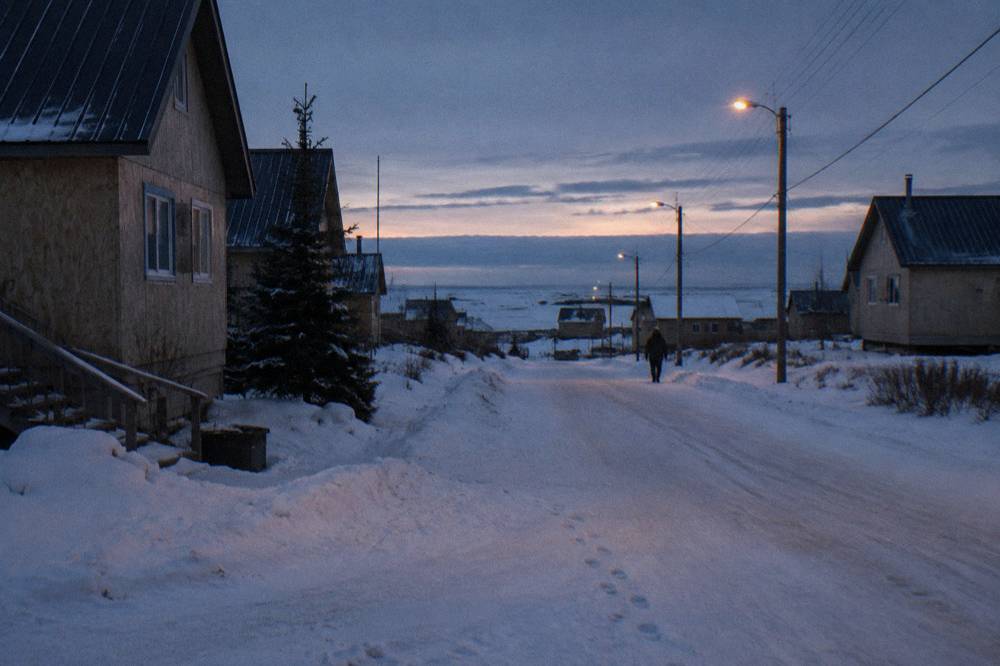A small northern town street at dusk with streetlights glowing and snow on the ground