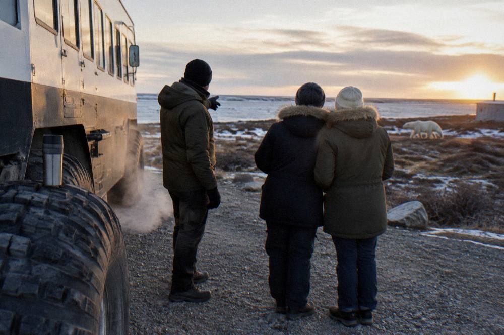 A guide in cold-weather gear standing beside a large tundra vehicle speaking with a small group of guests