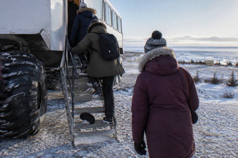 A small group of travelers in winter clothing boarding a large wheeled tundra vehicle at a launch site