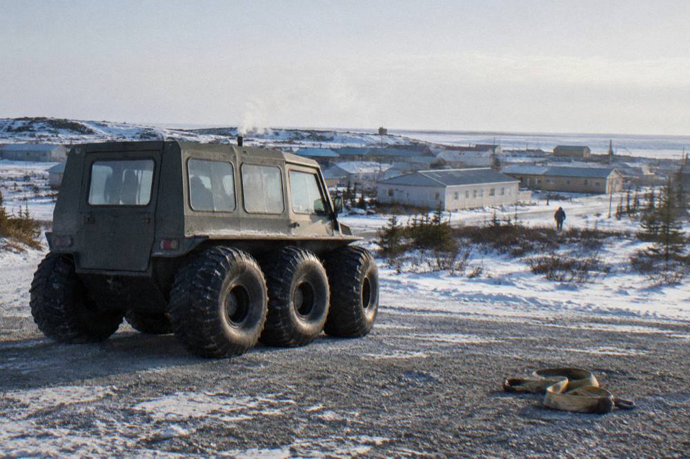 A heated tundra vehicle with large tires parked on gravel near a northern town