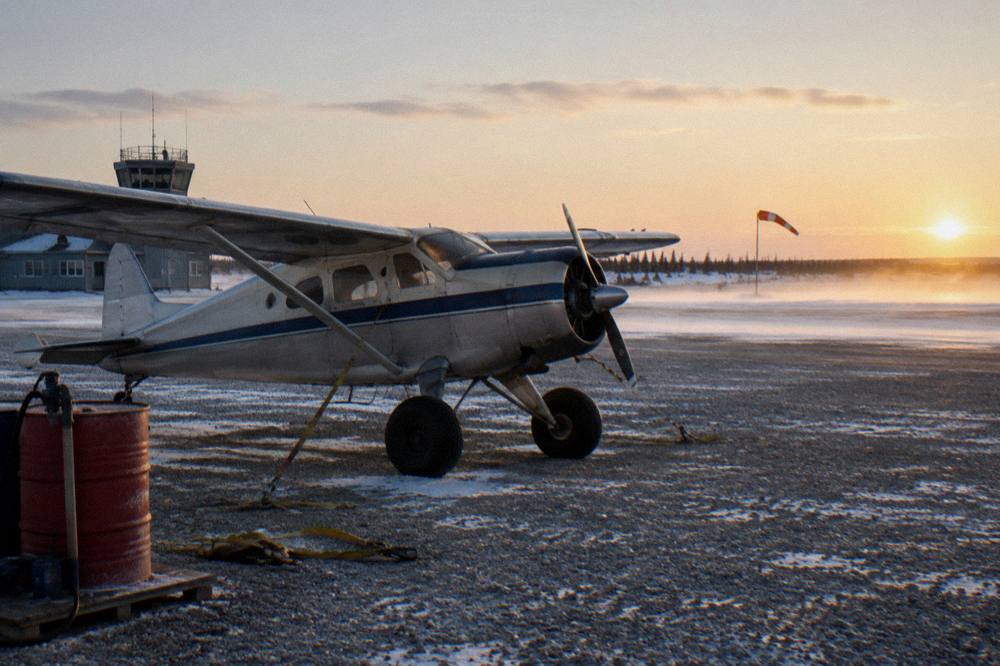 A small aircraft parked on a gravel runway beside a remote northern lodge building